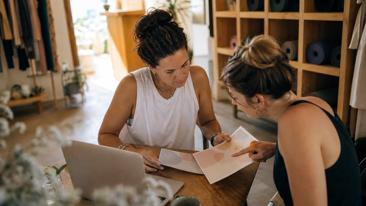 Yoga studio owner and her manager at a small table comparing two printed software quotes beside a laptop