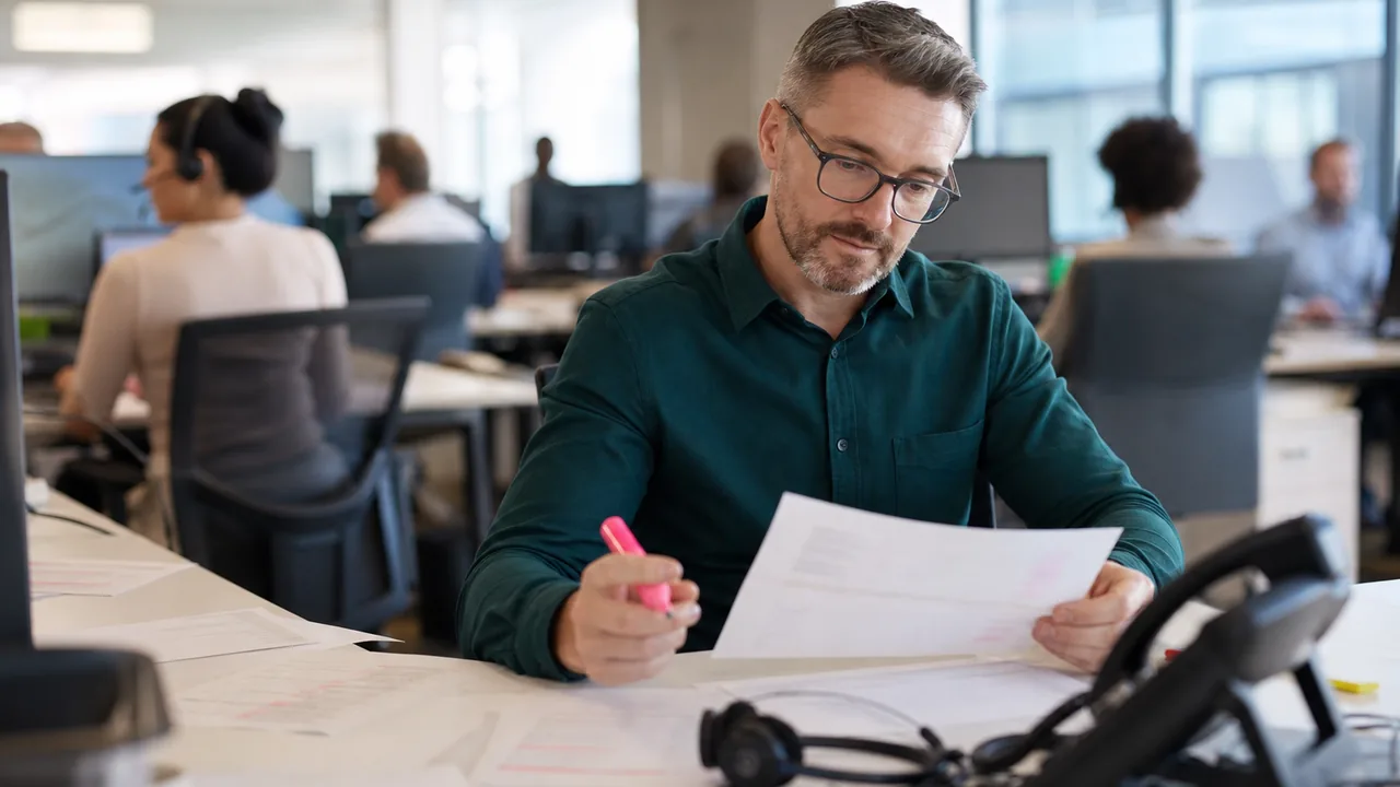 A retail customer support manager reviewing printed contact center usage invoices at a desk beside a desk phone and headset